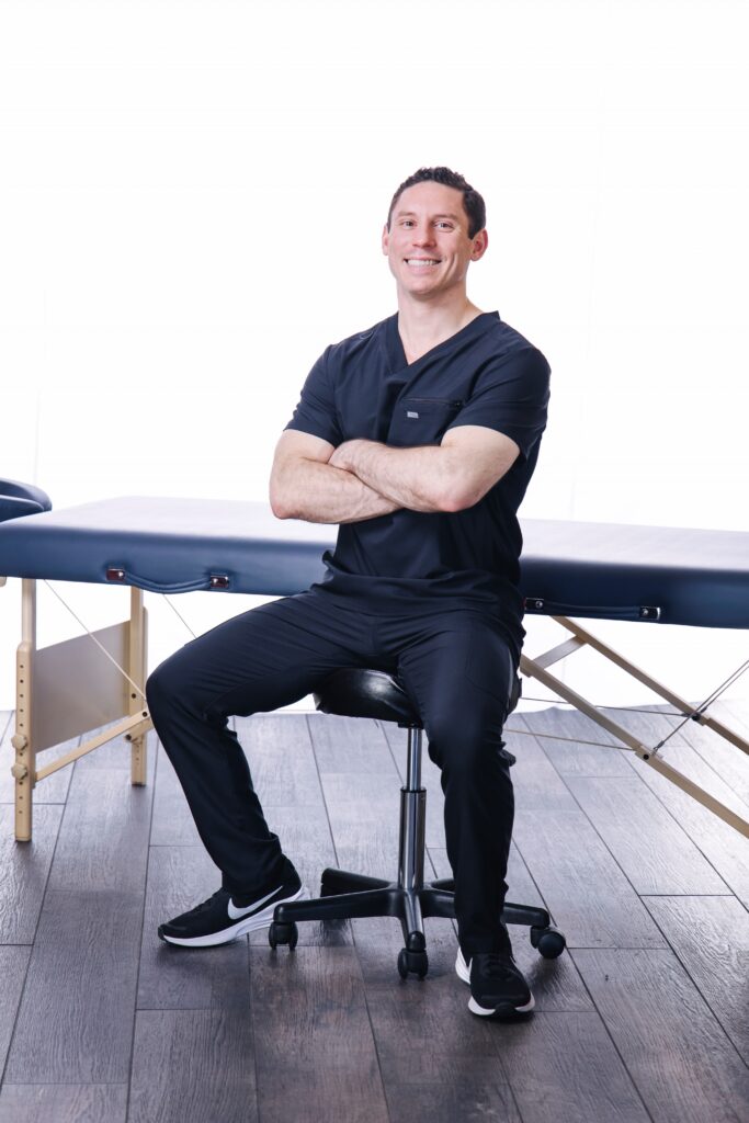 Ben, licensed massage therapist, seated beside his massage table